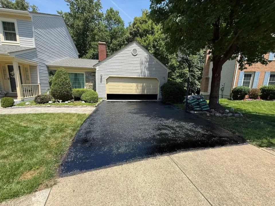 A freshly sealed black asphalt driveway leading to a suburban house with an open garage.
