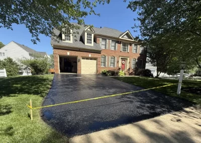 A freshly paved asphalt driveway in front of a brick suburban house, blocked off by yellow caution tape.
