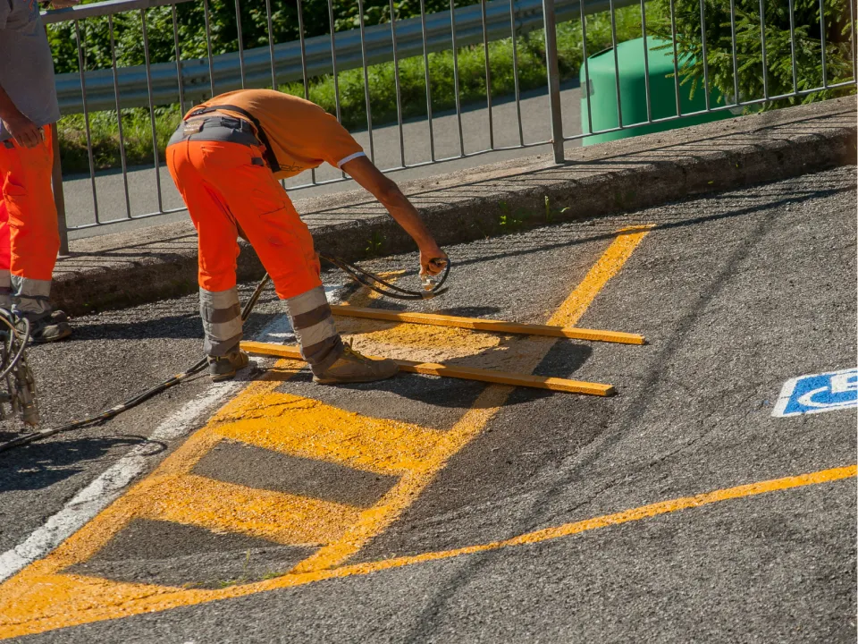 A worker in orange high-visibility clothing paints yellow parking lines on asphalt.