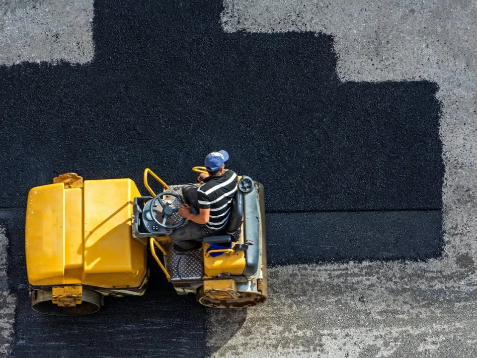 A person operating a yellow road roller to pave a patch of fresh asphalt.