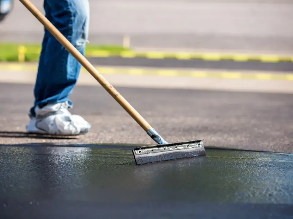 A person using a squeegee to spread asphalt sealant on a driveway.