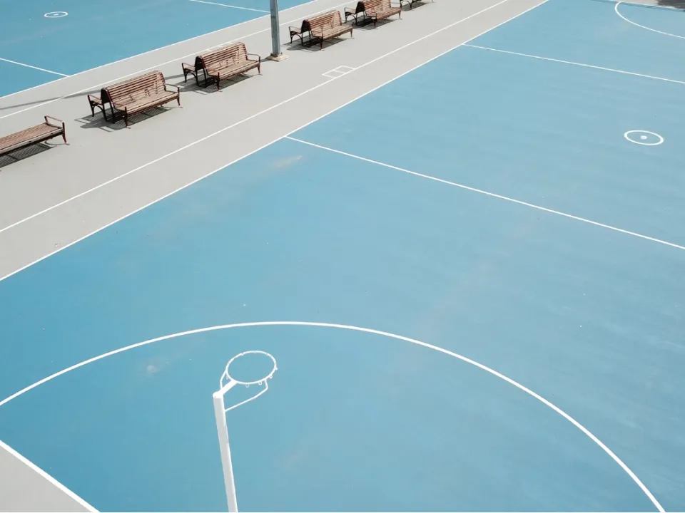 An outdoor blue netball court with a row of wooden benches along the side.